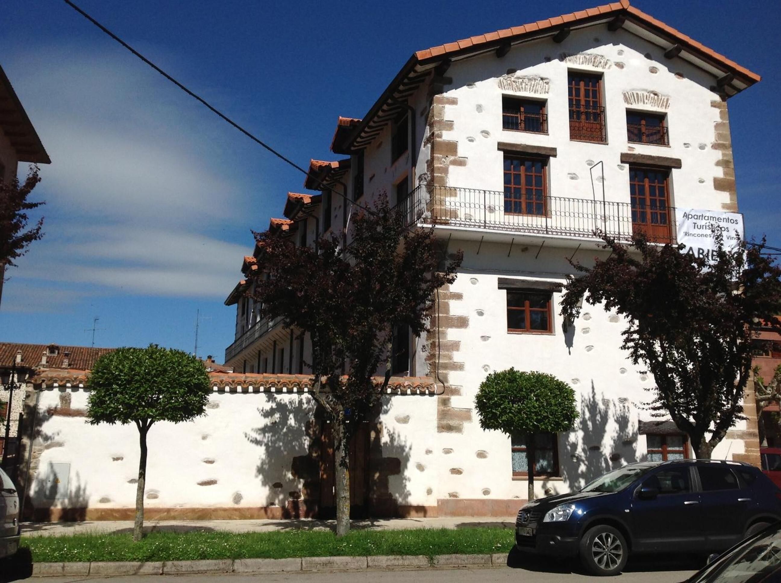 A two-story building with a white facade and brown roof tiles. There are trees in front of the building, and a car is parked on the street.