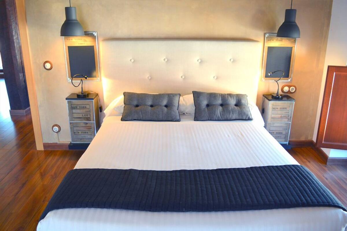 A large bed with a tufted headboard, two bedside tables with lamps, and a dark throw blanket.