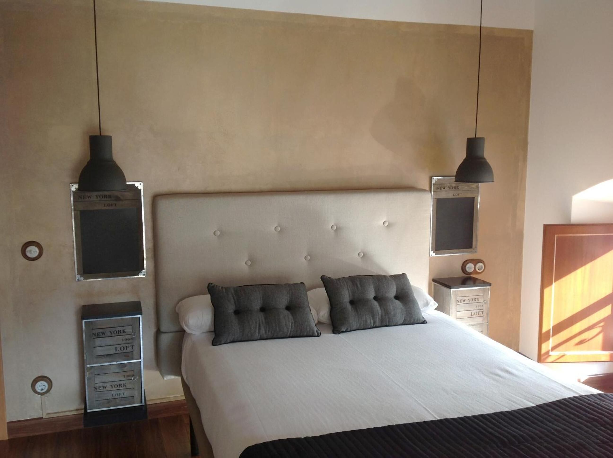 Un dormitorio con cabecera capitonada, dos mesitas de noche, una cama con ropa blanca y dos almohadas grises, y dos lámparas colgantes.