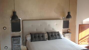 Un dormitorio con cabecera capitonada, dos mesitas de noche, una cama con ropa blanca y dos almohadas grises, y dos lámparas colgantes.