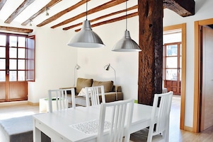 Comedor con mesa y sillas blancas, dos lámparas colgantes, techo de madera y sofá con cojines.