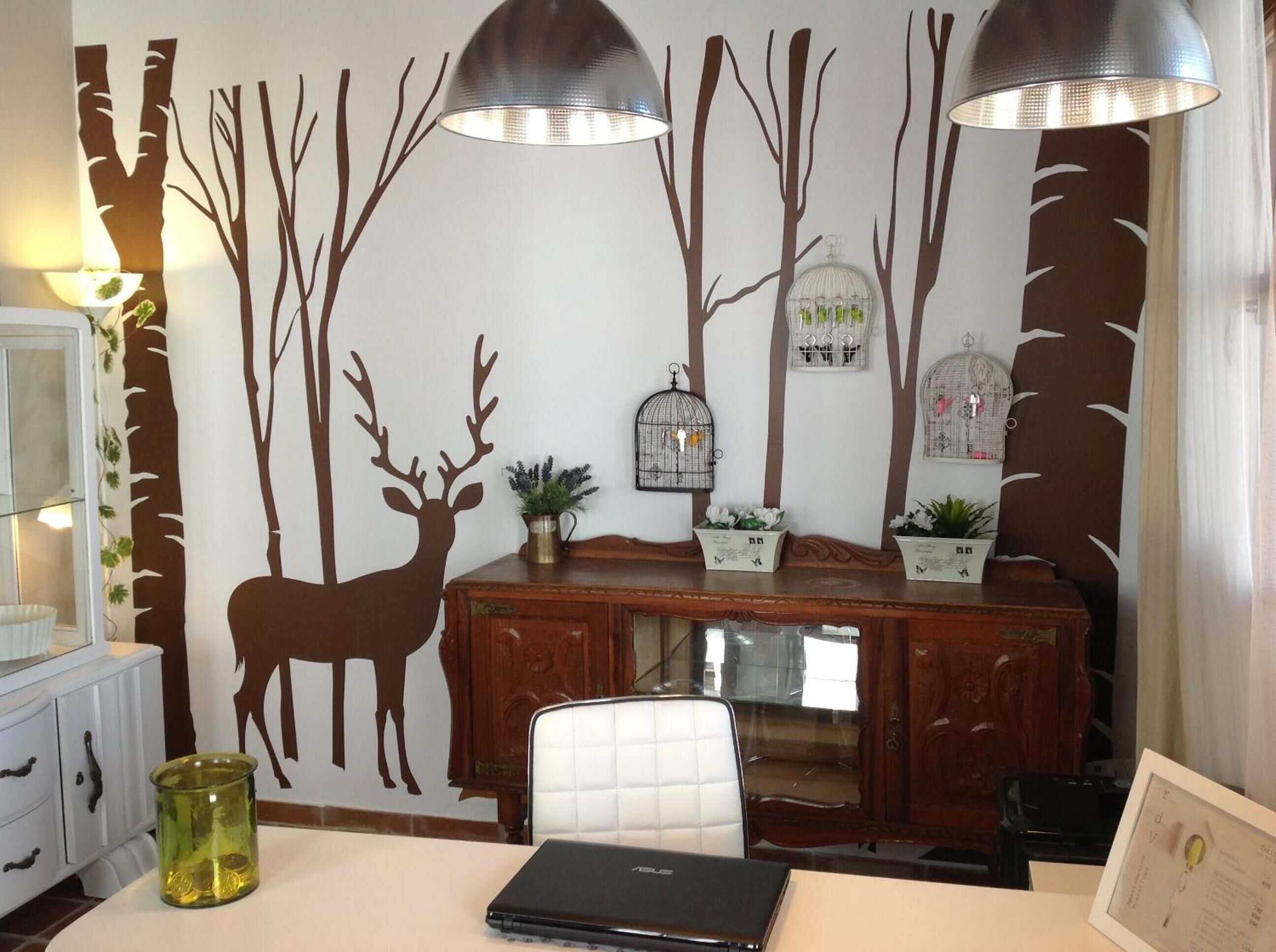 Un comedor con una mesa de madera, sillas y una computadora portátil. Paneles decorativos en la pared con siluetas de árboles y ciervos.
