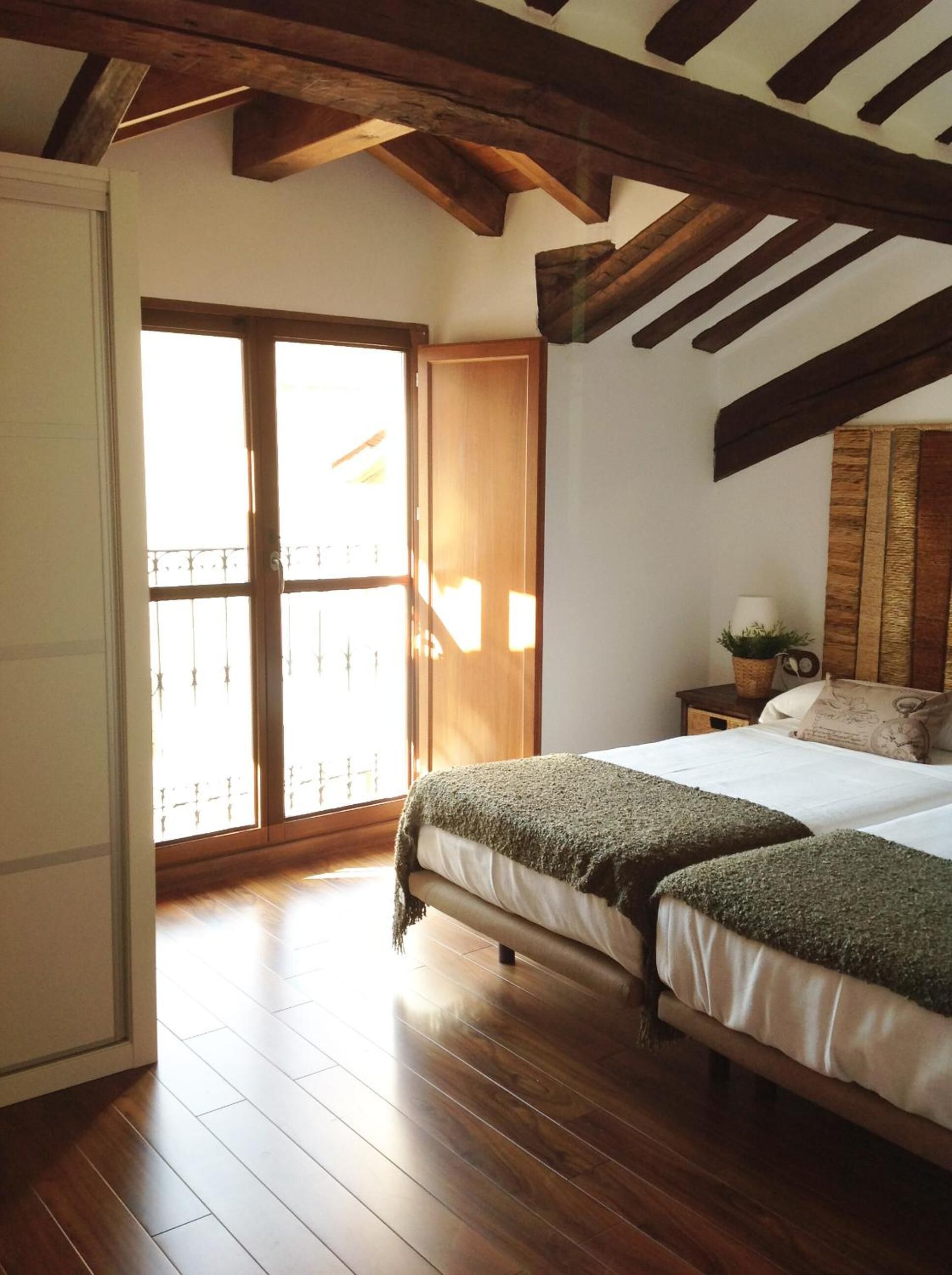 Un dormitorio con piso de madera, una cama con manta verde, una mesita de noche con una planta y vistas al exterior mediante una puerta corredera.