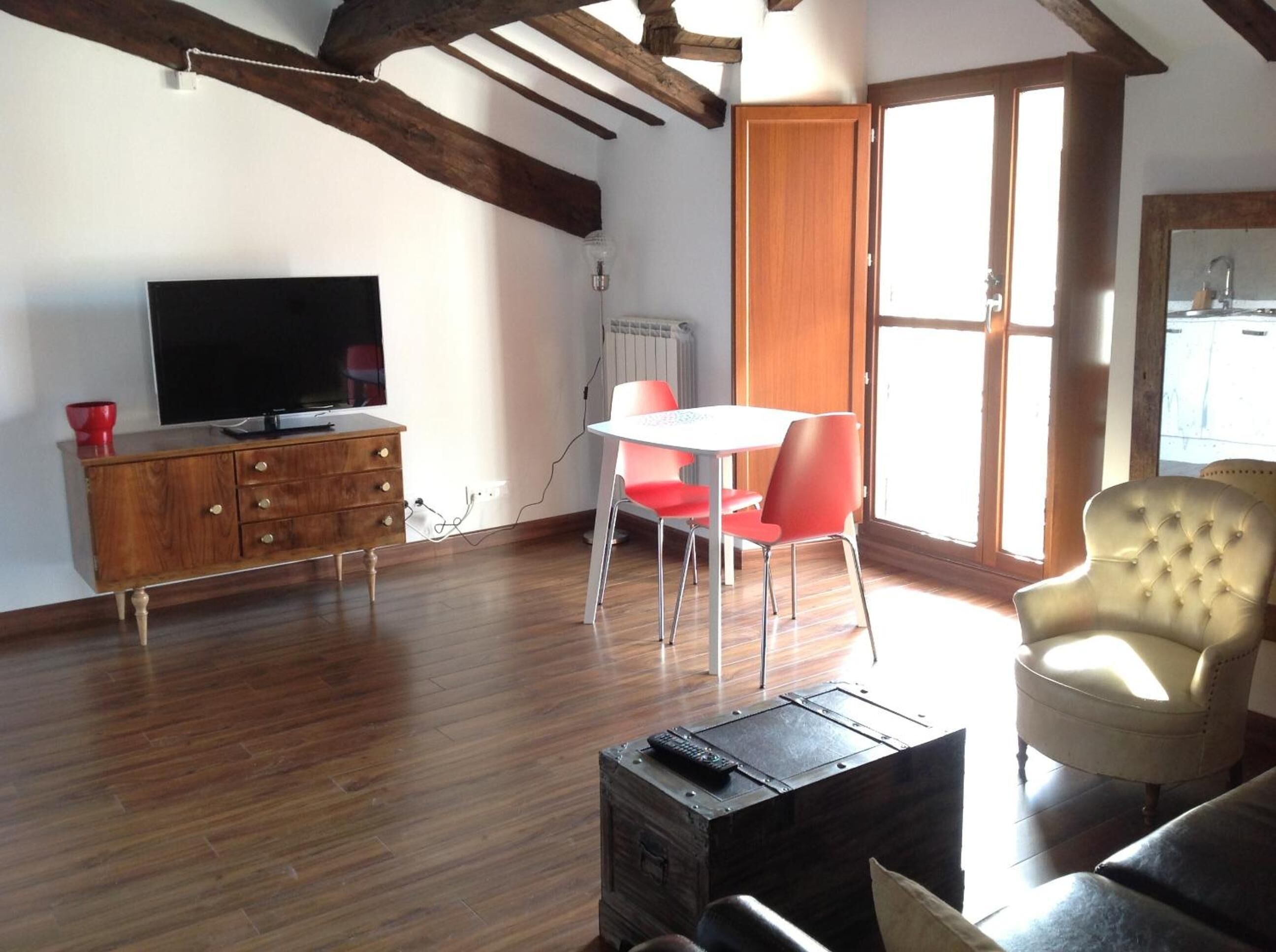 A living room with a wooden floor, a TV on a wooden cabinet, a dining table with red chairs, and a large window with a view.