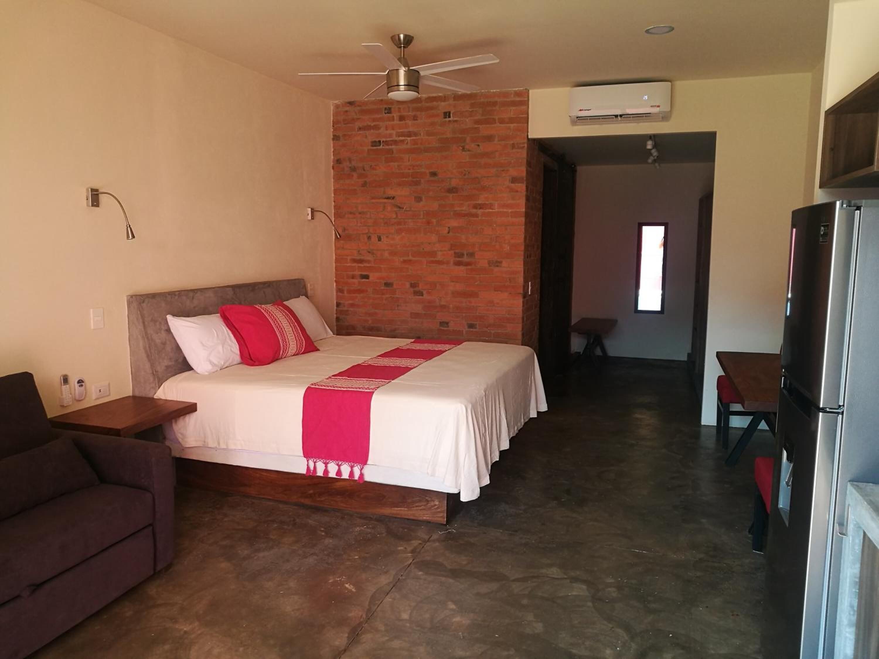 Una habitación de hotel con una cama, ventilador de techo, pared de ladrillo y kitchenette.