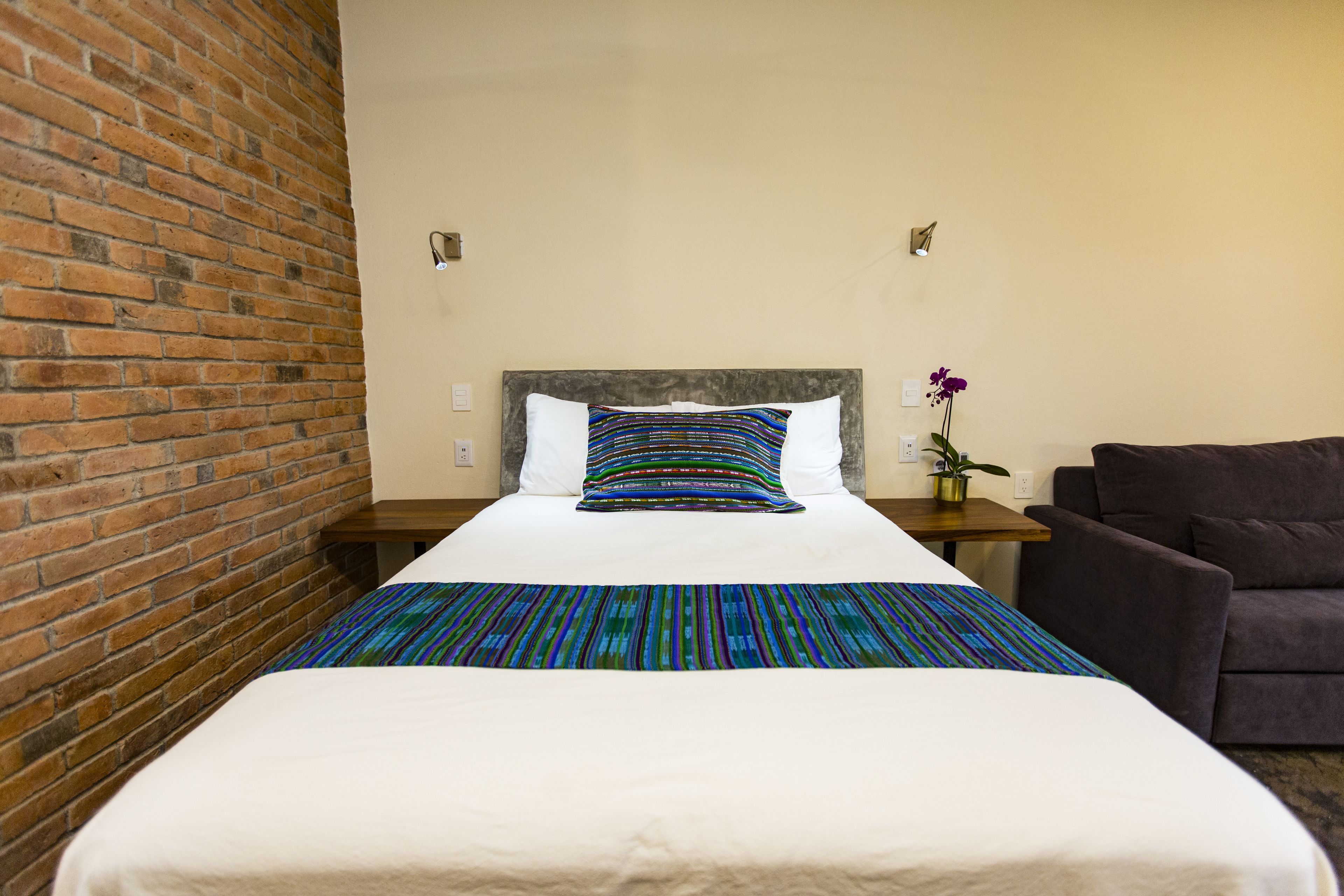 A hotel room with a large bed, a brick wall, a gray headboard, a small table with a potted plant, and a dark-colored armchair.