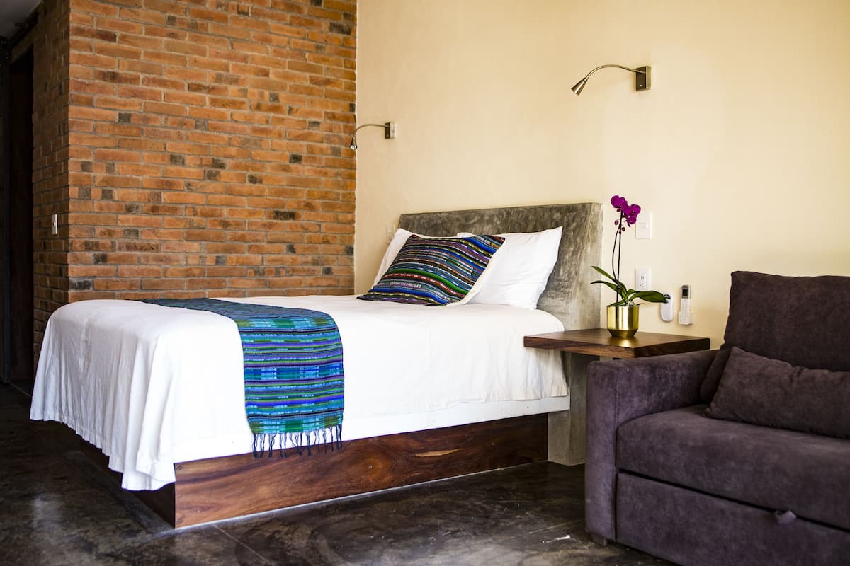 A neatly made bed with a white comforter and a colorful blanket, a wooden nightstand with a vase of flowers, and a dark-colored sofa.
