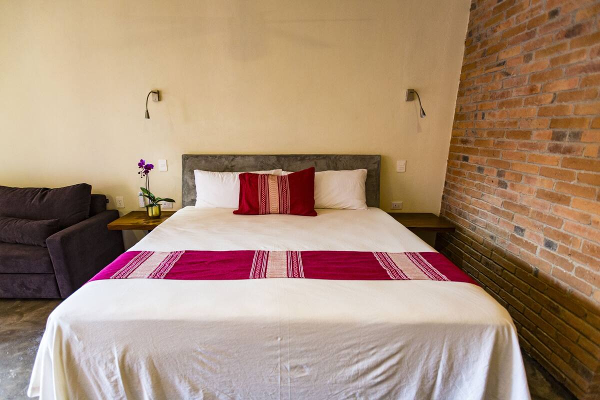 A neatly made bed with a white and red bedspread, a dark brown headboard, and a brick wall to one side.