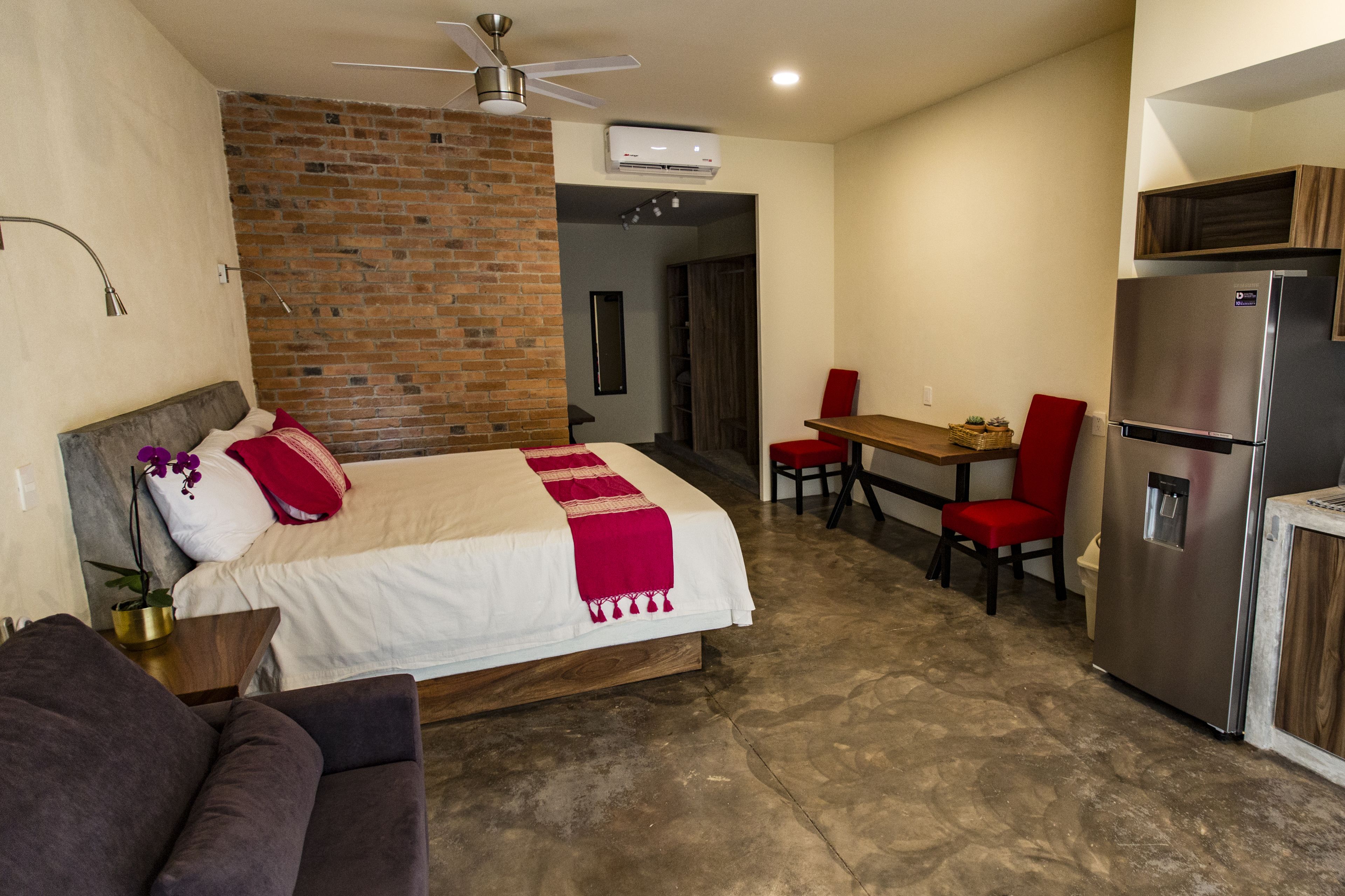 A hotel room with a bed, a small seating area, a refrigerator, and a brick wall.