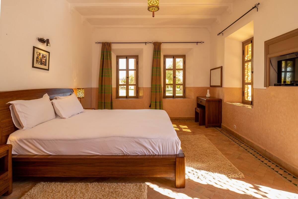 Une chambre à coucher avec un lit en bois, deux fenêtres avec des rideaux, une table de chevet, une commode, une lampe et un tableau au mur.