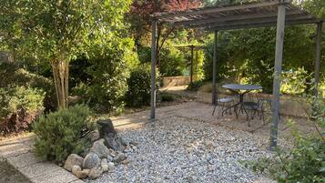 A covered patio area with a table and chairs, surrounded by a gravel path and various plants.