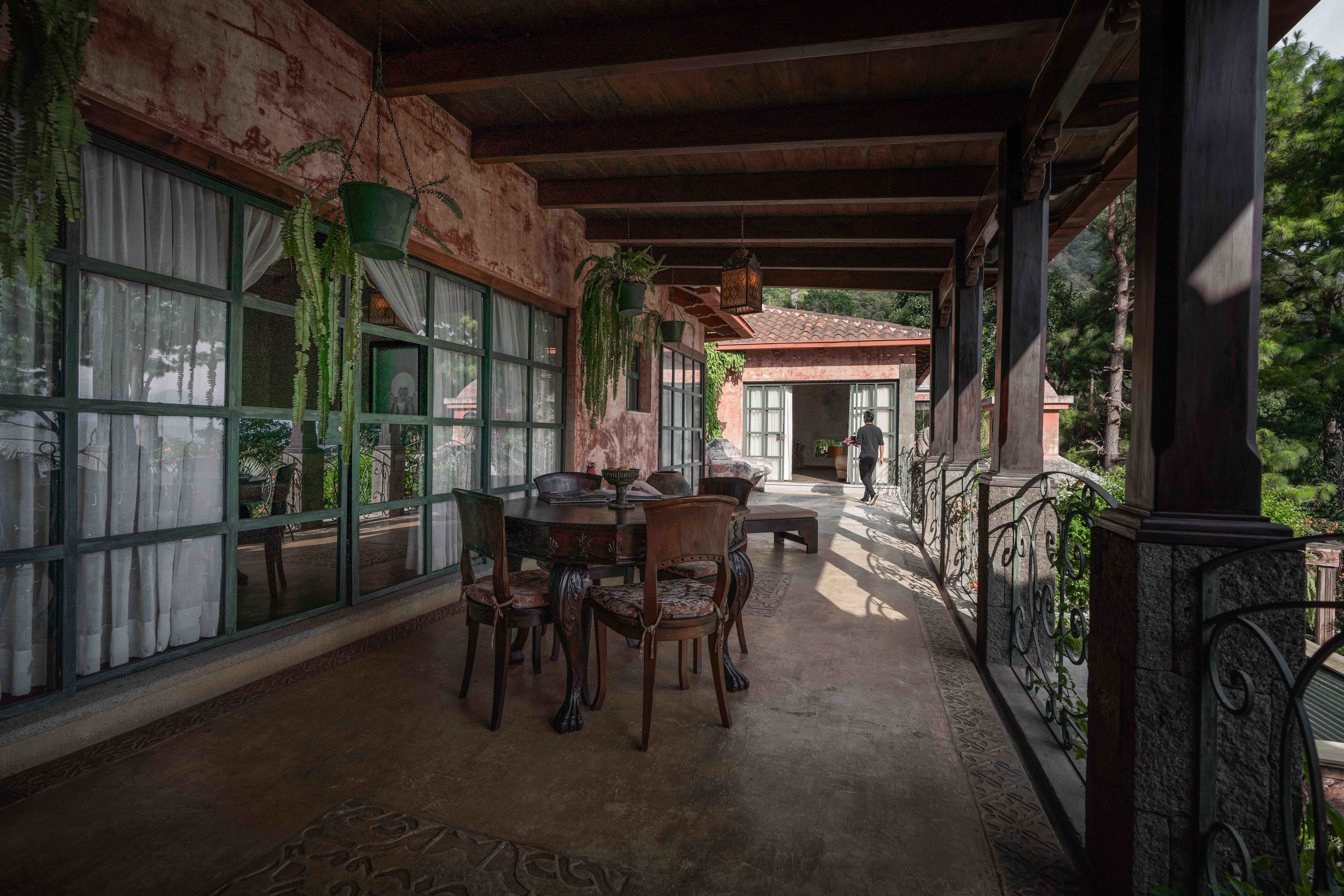 Un patio cubierto con una mesa y sillas de comedor, ventanas grandes con cortinas, plantas colgantes y una vista de un edificio al fondo.