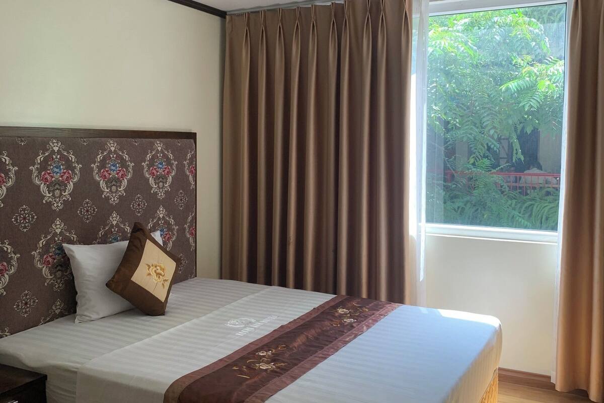 A hotel room with a large bed, a headboard with a floral pattern, a window with curtains, and a view of greenery outside.