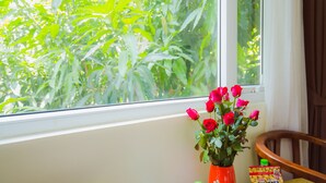 A window with a view of green foliage, a vase with red roses, and a chair. - Hoang Anh 1 Hotel (Hanoi)