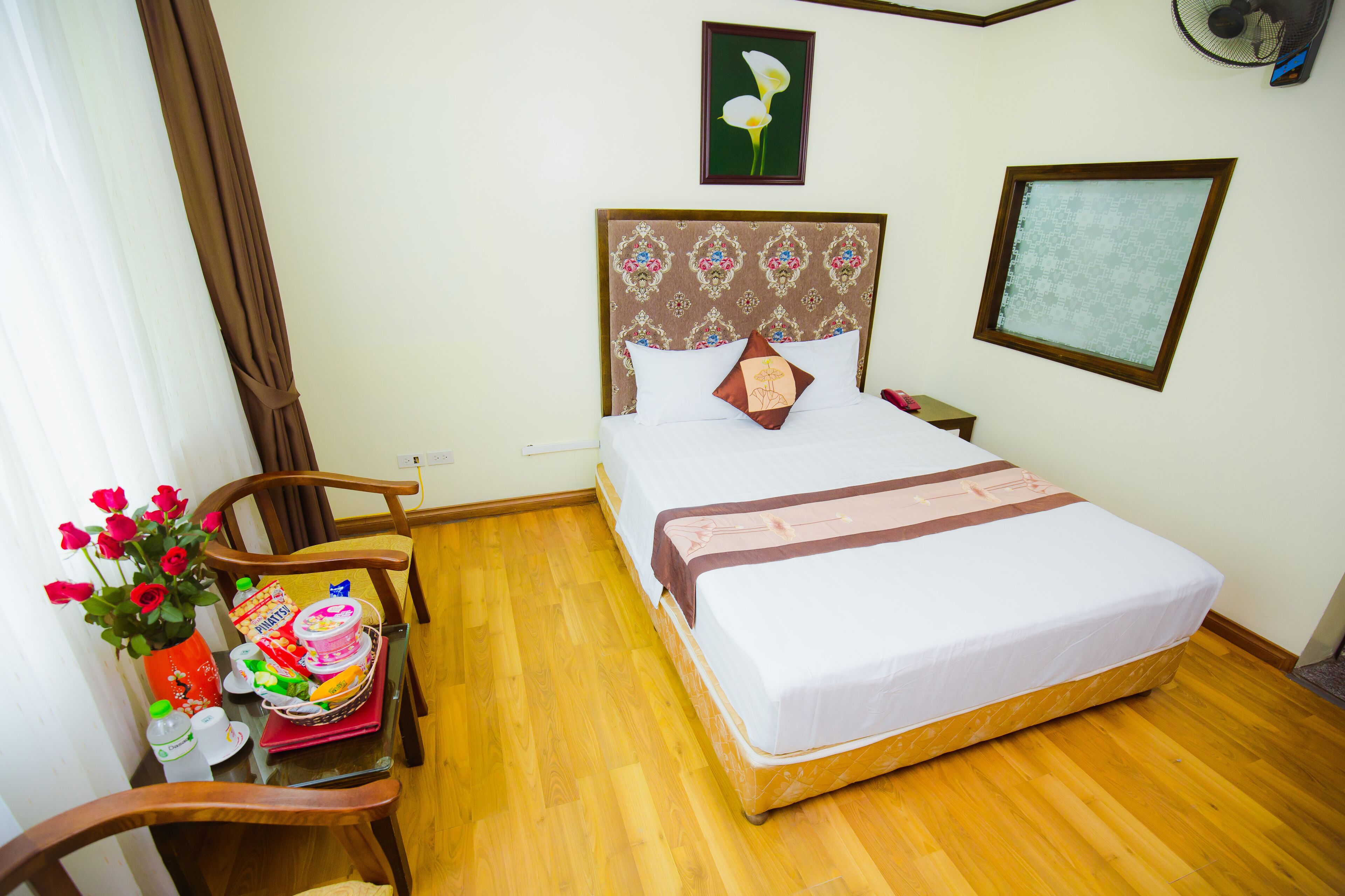 A neatly made bed with a patterned headboard, a bedside table with a vase of flowers, and a wooden floor.