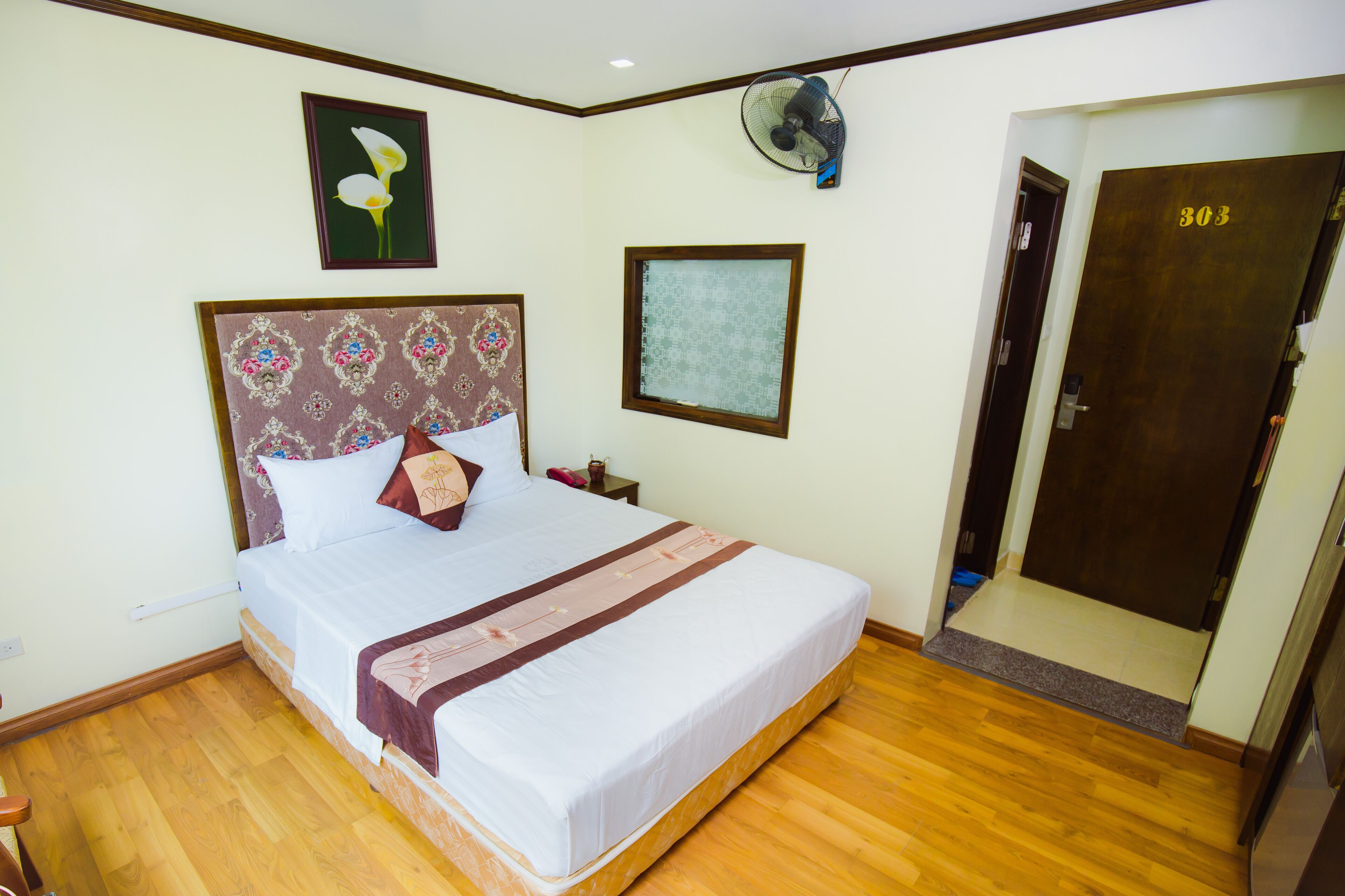 Una habitación de hotel con una cama grande, piso de madera, una puerta con el número 303 y un ventilador de pared.