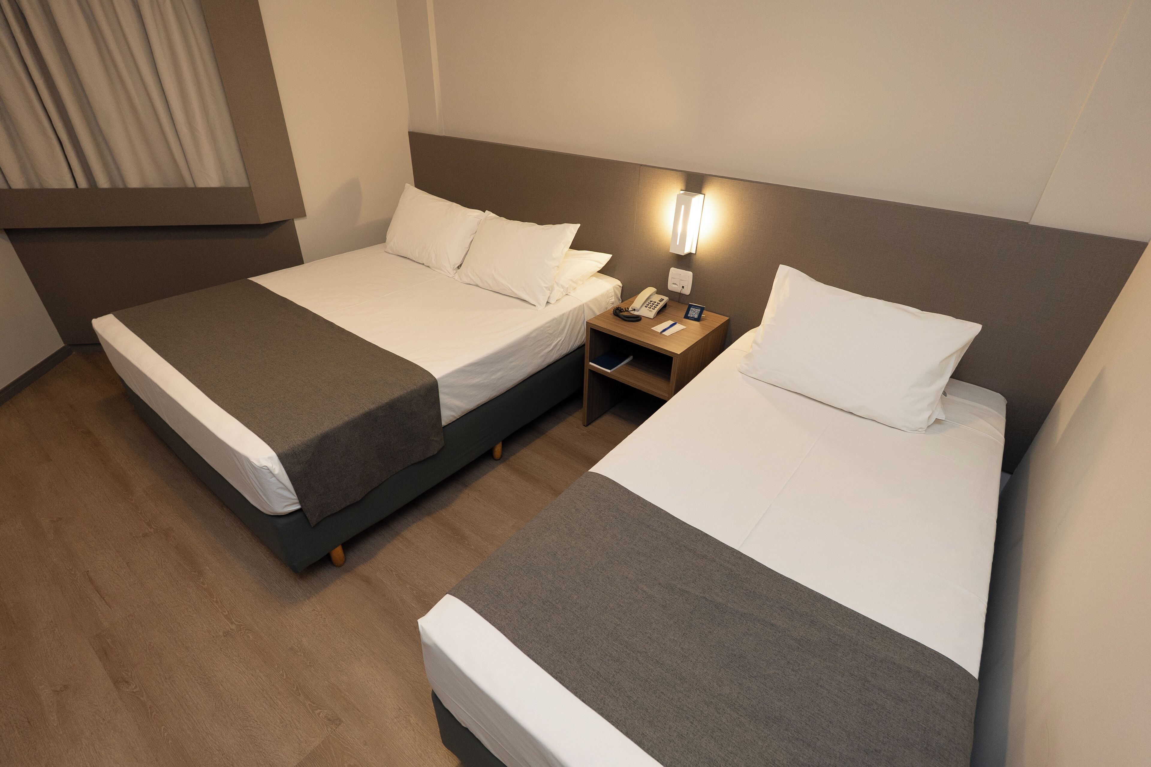 Deux lits simples avec une literie grise et blanche, une table de chevet avec un téléphone et une lampe, et un plancher en bois.