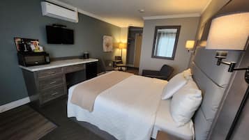 A hotel room with a bed, bedside table, desk, chair, and an air conditioning unit.