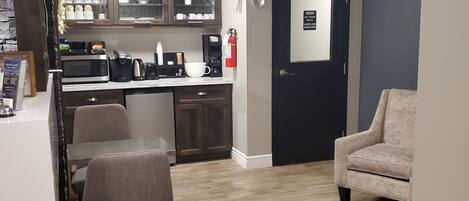 A modern living room with a grey sofa, a dining area with a table and chairs, a kitchen area with appliances and cabinets, and a door with a fire safety sign.