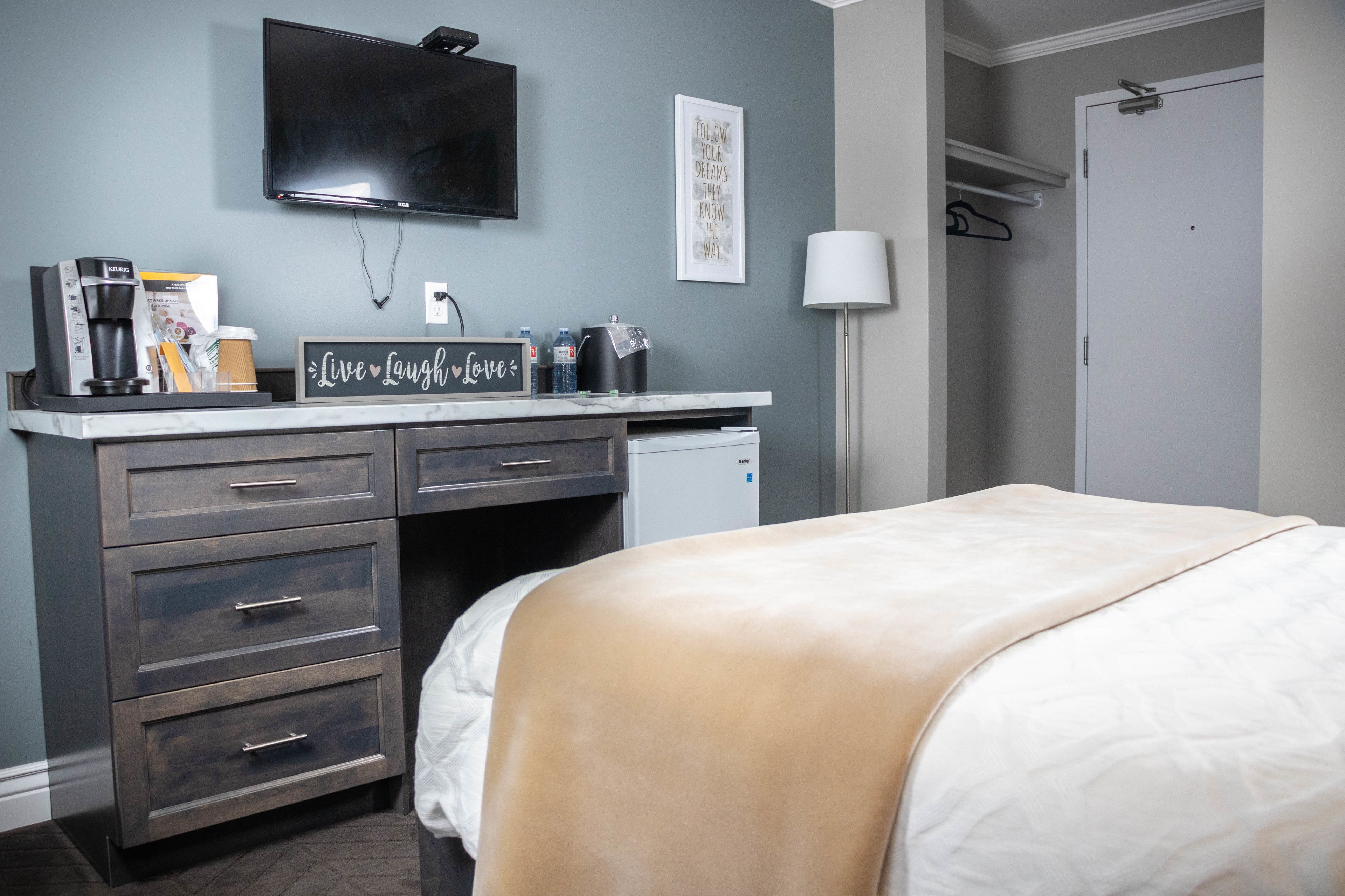 A hotel room with a bed, a desk with drawers, a coffee maker, a flat-screen TV, and a wall-mounted lamp.