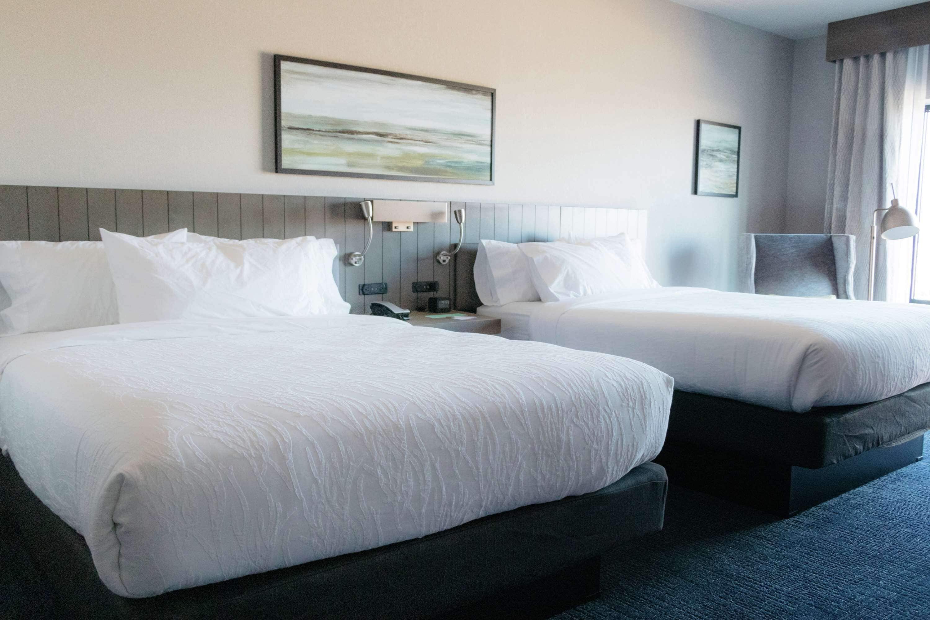A hotel room with two beds, a headboard with a light switch, a bedside table, and a wall-mounted artwork.