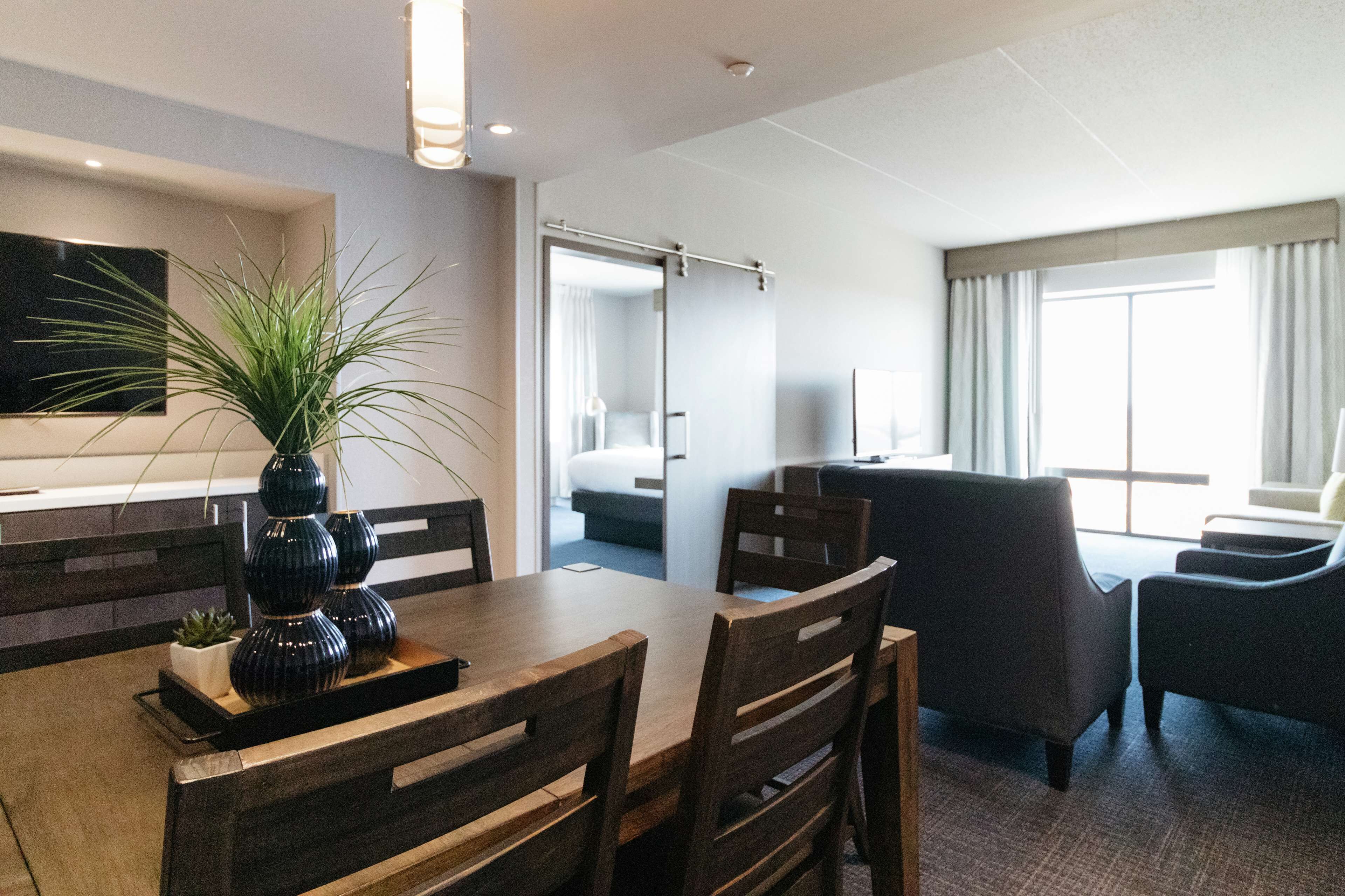 Habitación de hotel con zona de comedor, un ventanal con cortinas, una cama y un televisor instalado en la pared.
