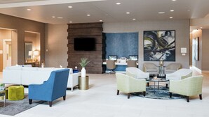 A modern lobby with a white sofa, blue armchair, and green armchairs. There is a fireplace, a desk with two monitors, and a large abstract painting on the wall. - Hilton Garden Inn Madison Sun Prairie (Sun Prairie)