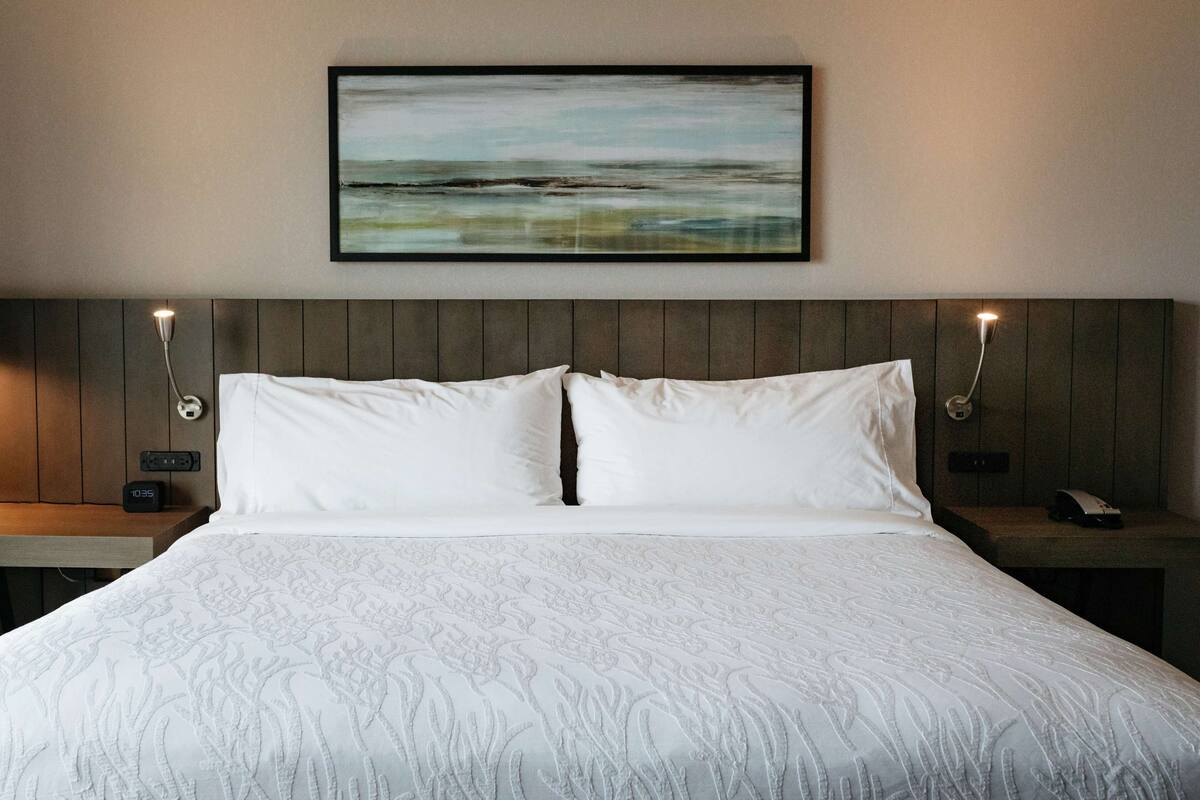 A neatly made bed with a patterned bedspread, a framed abstract painting above it, and two bedside tables with lamps and a clock on the left side.