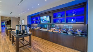 A hotel reception area with a coffee station, a TV screen displaying a news program, and a counter with various food items. - Hilton Garden Inn Madison Sun Prairie (Sun Prairie)