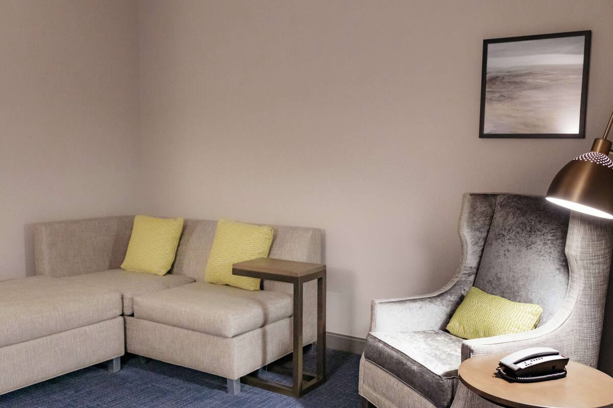 A modern hotel room with a sectional sofa, a small side table, a large armchair, and a lamp.