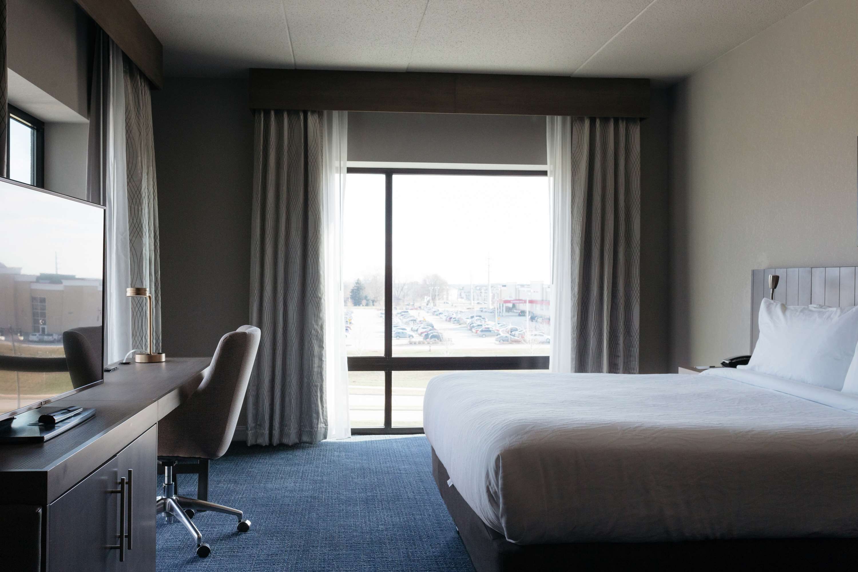 A hotel room with a bed, a desk, a chair, and a large window with curtains.