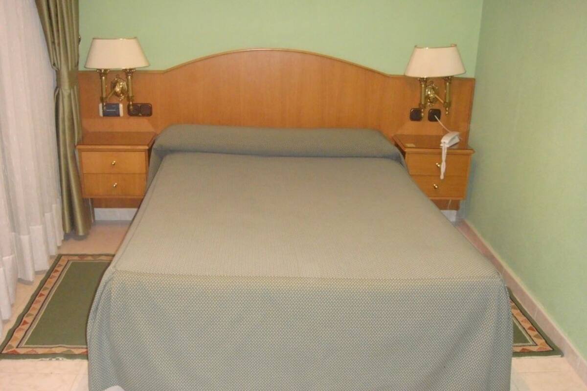 Una cama individual con cabecera de madera, dos mesitas de noche con lámparas, un teléfono y una colcha verde.