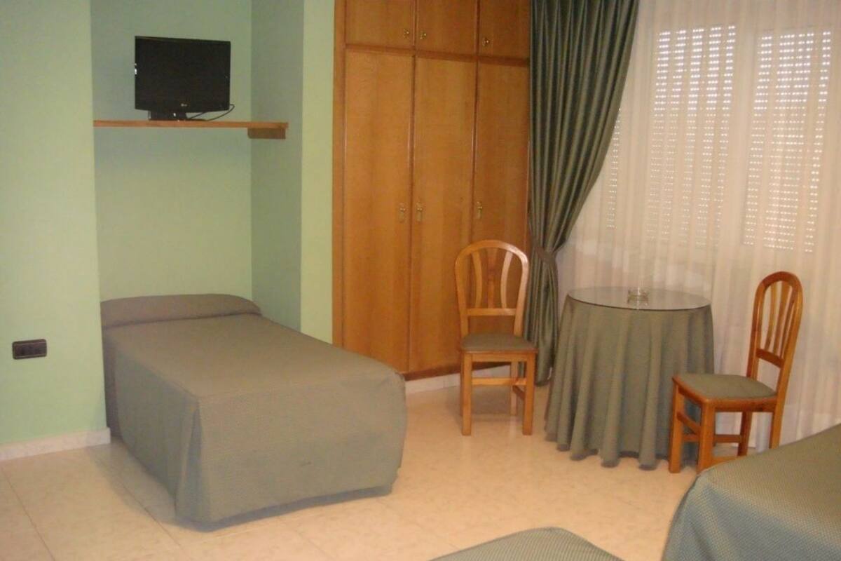 Una habitación con una cama, una mesita pequeña, dos sillas, un televisor en un estante y un armario.
