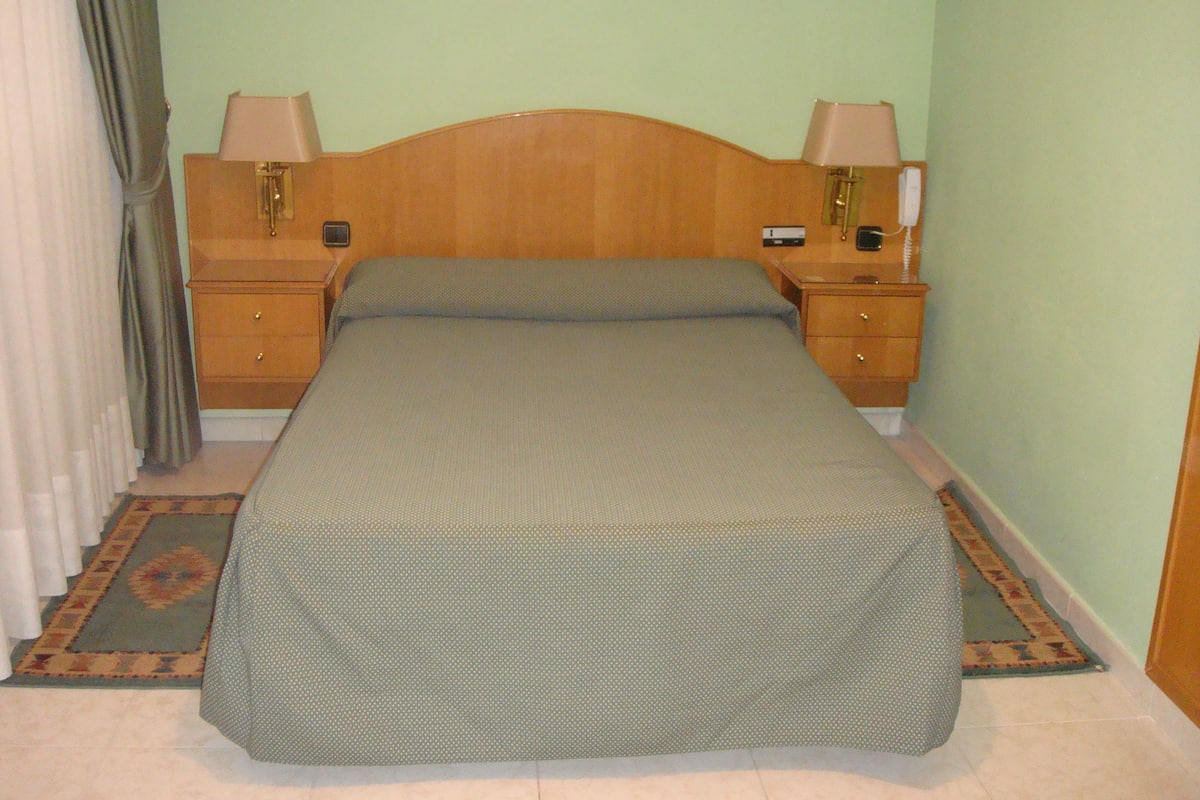 Una cama individual con cabecera de madera, dos mesitas de noche con lámparas, una colcha estampada y una alfombra decorativa.