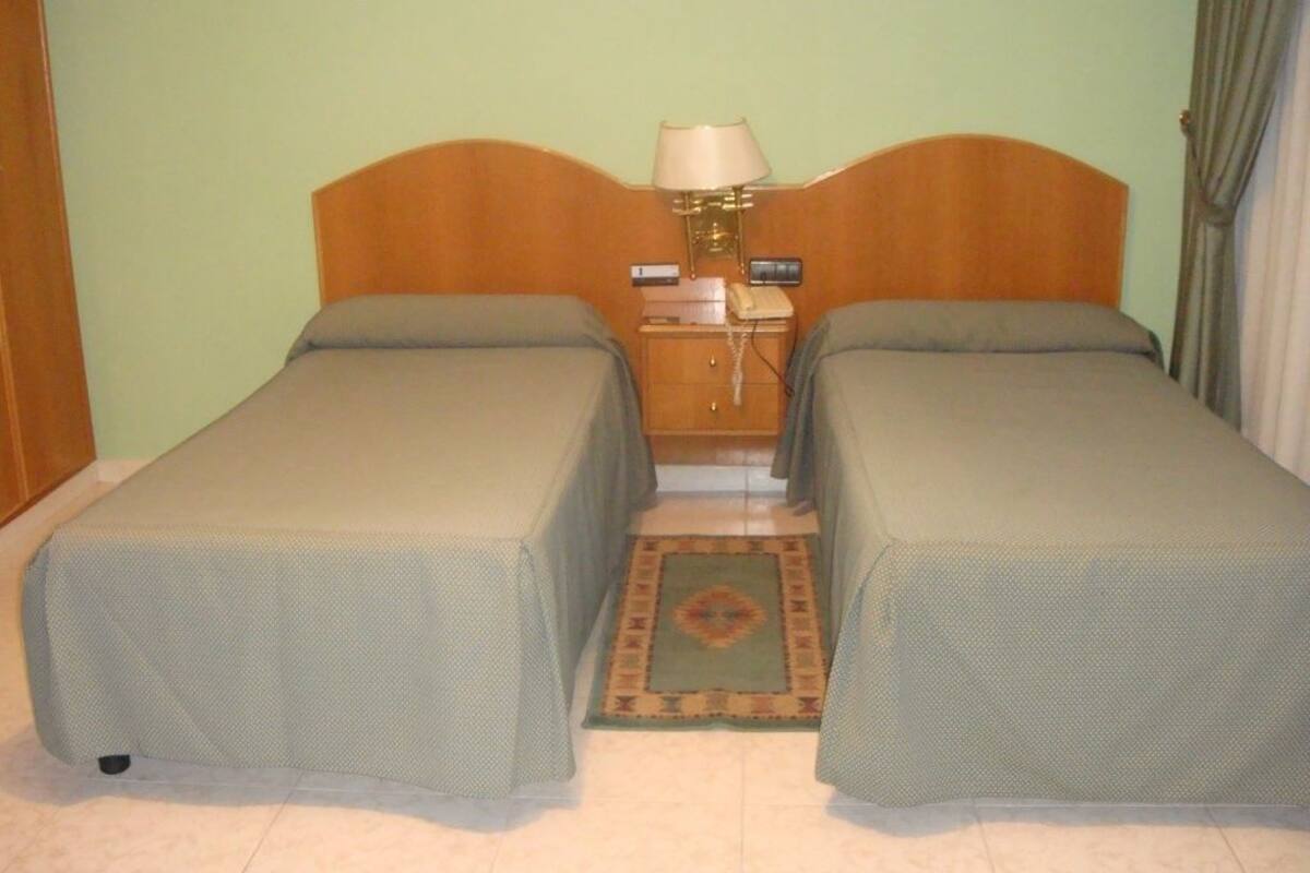 Habitación doble con dos camas individuales, una mesita de noche con lámpara, teléfono y una pequeña mesa.
