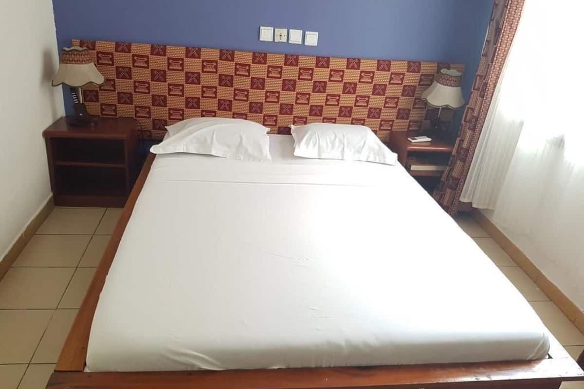 Una cama bien hecha en una habitación con un cabecero estampado, una mesita de noche con lámpara y una ventana con cortinas.