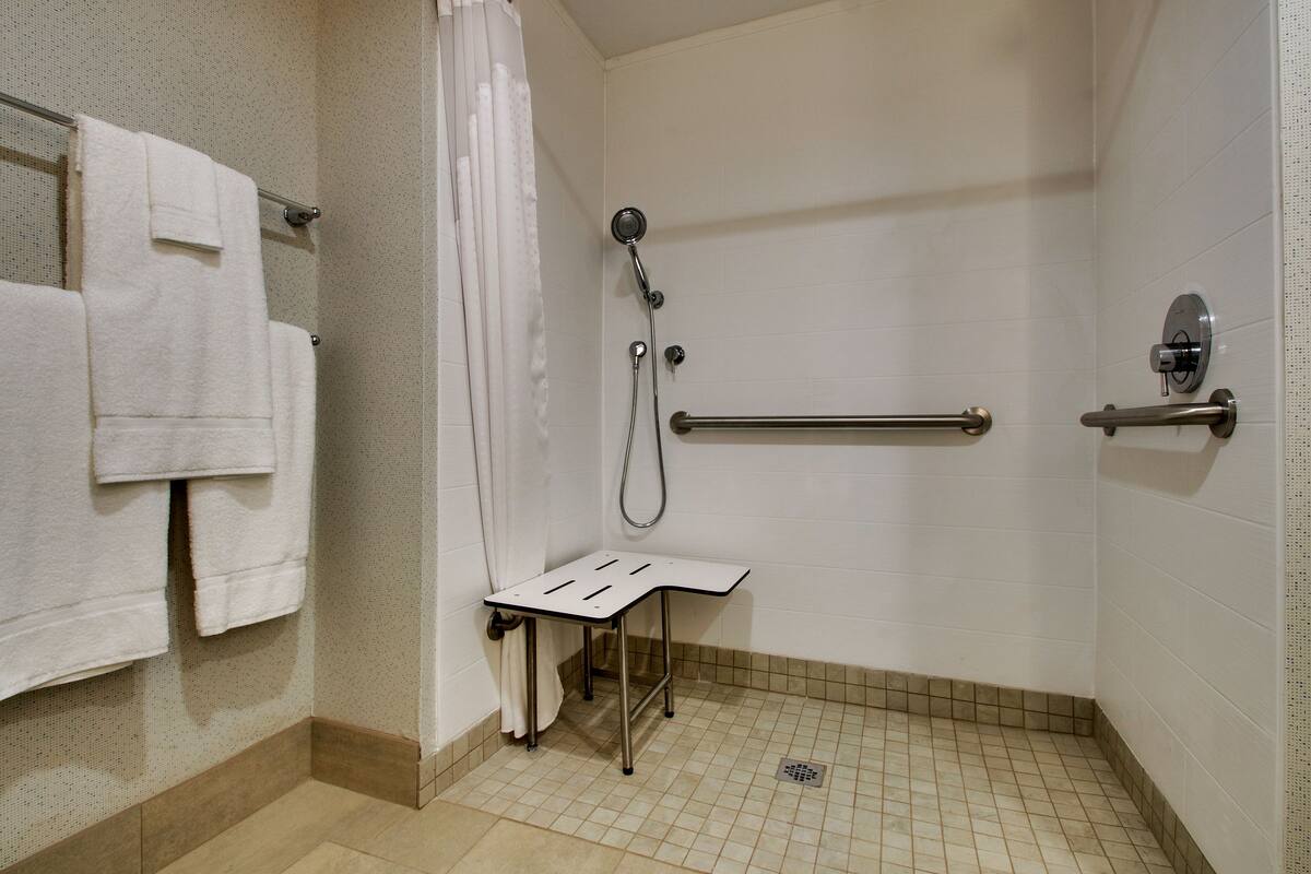 A wheelchair-accessible shower with grab bars, a white tiled floor, and a towel rack with towels.