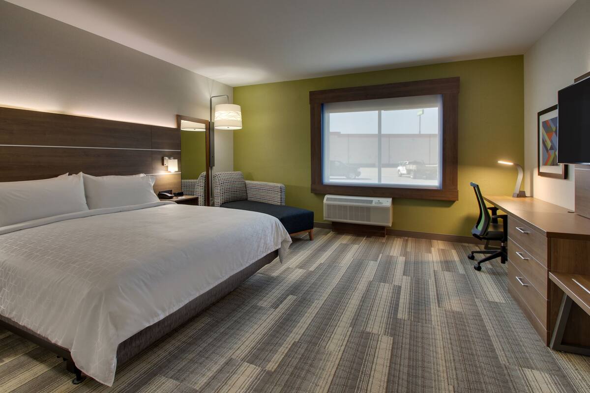 A hotel room with a large bed, a desk with a chair, a window with a view of the outside, and a wall-mounted TV.