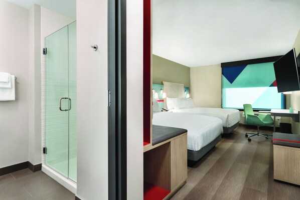 Ein Hotelzimmer mit Duschkabine aus Glas, zwei Betten, einem Schreibtisch und einem Stuhl.