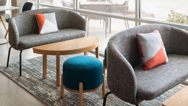 Un interior moderno con sillones grises, una mesa de madera y un reposapiés azul. Amplias ventanas ofrecen vistas al exterior.