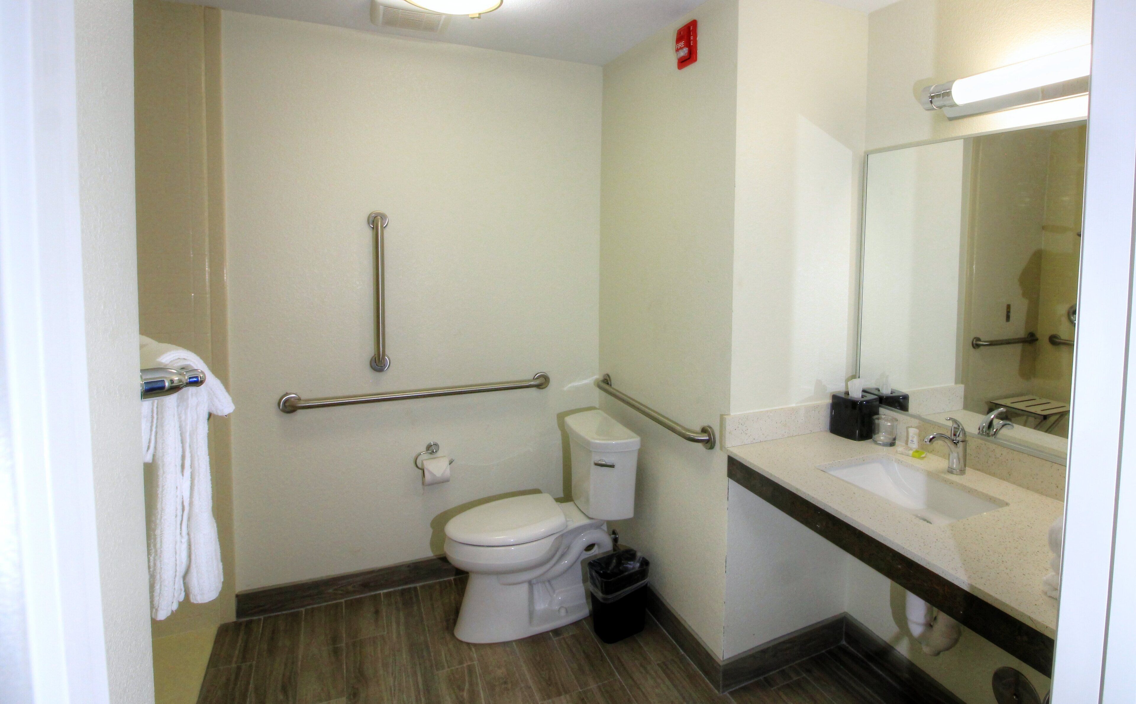 Une salle de bain équipée de toilettes, de barres d’appui, d’un lavabo, d’un miroir et d’une poubelle.