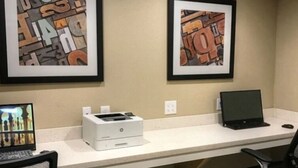 A workspace with two framed abstract artworks, a desktop printer, a laptop, and a monitor displaying a cityscape. - Candlewood Suites Memphis East by IHG (Memphis)