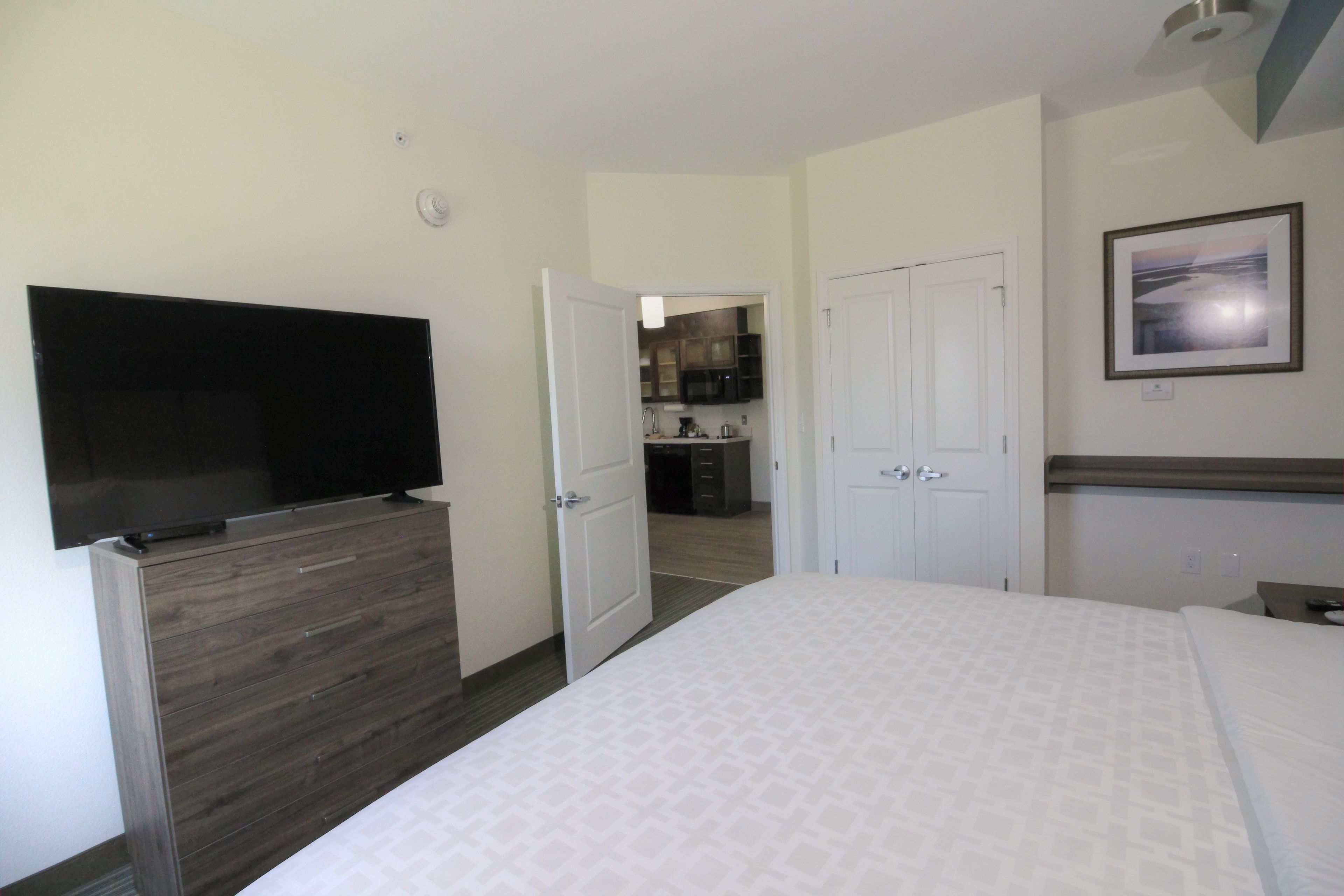 Habitación de hotel con una cama, un televisor en una cómoda, una puerta que da a una cocina y un cuadro en la pared.