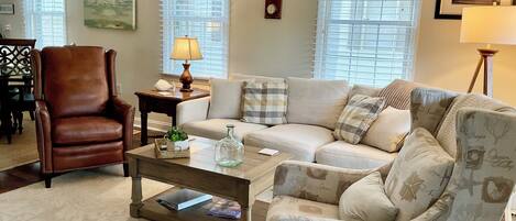 A living room with a brown leather chair, a beige sofa, and a patterned armchair. There is a coffee table with a lamp, a small side table, and a wall clock.