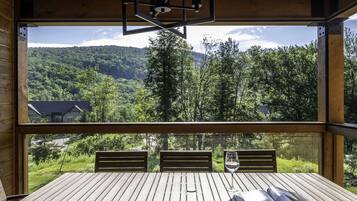 Une terrasse en bois avec une table et des chaises, donnant sur une vallée boisée, avec une montagne qui se profile à l’horizon.