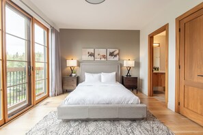 Ein Schlafzimmer mit einem großen Bett, zwei Nachttischen, einem Kopfteil und einem Fenster mit Vorhängen. Eine Tür führt in einen anderen Raum.
