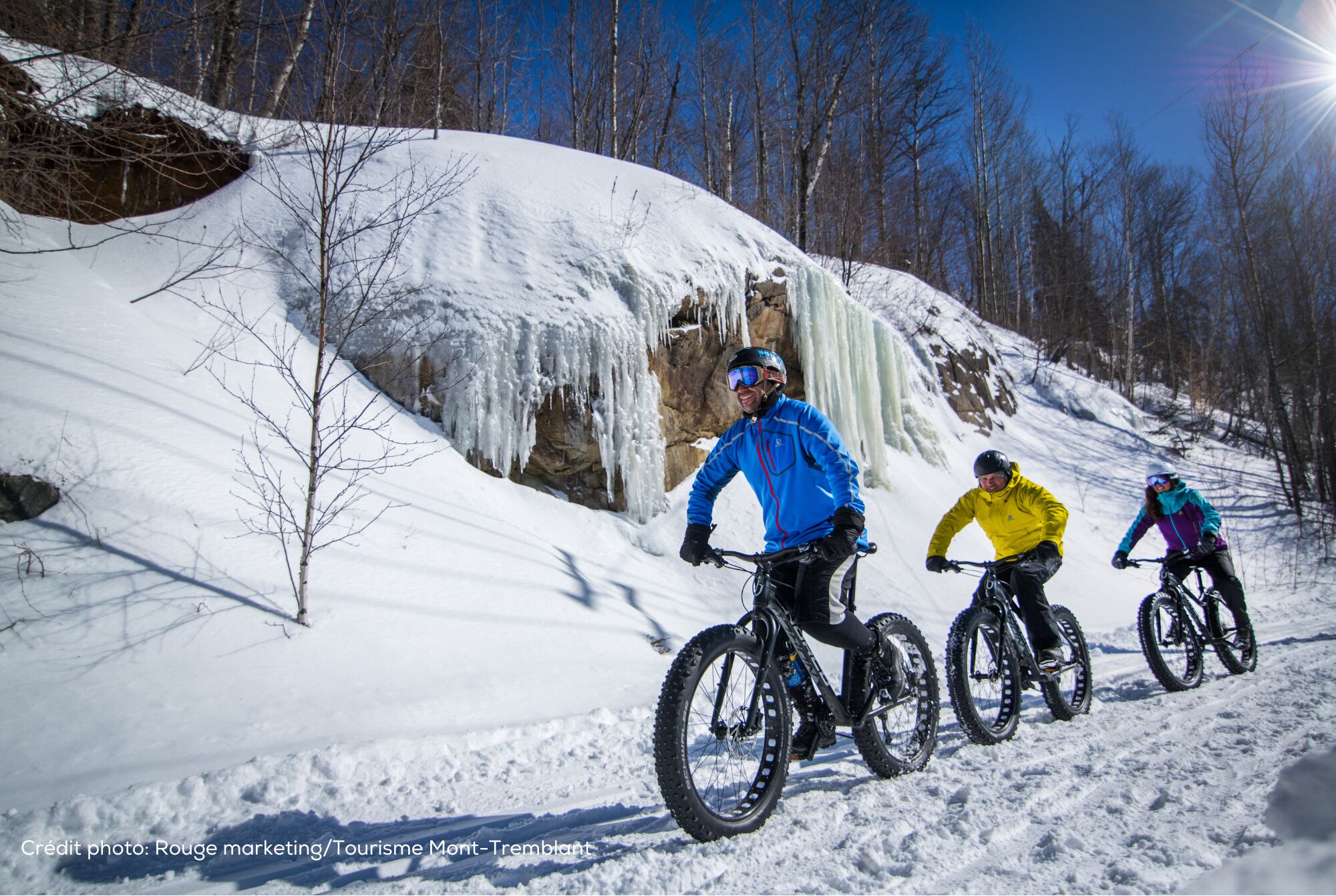 Three people on snow bikes on a snowy slope with a rocky cliff and icicles.