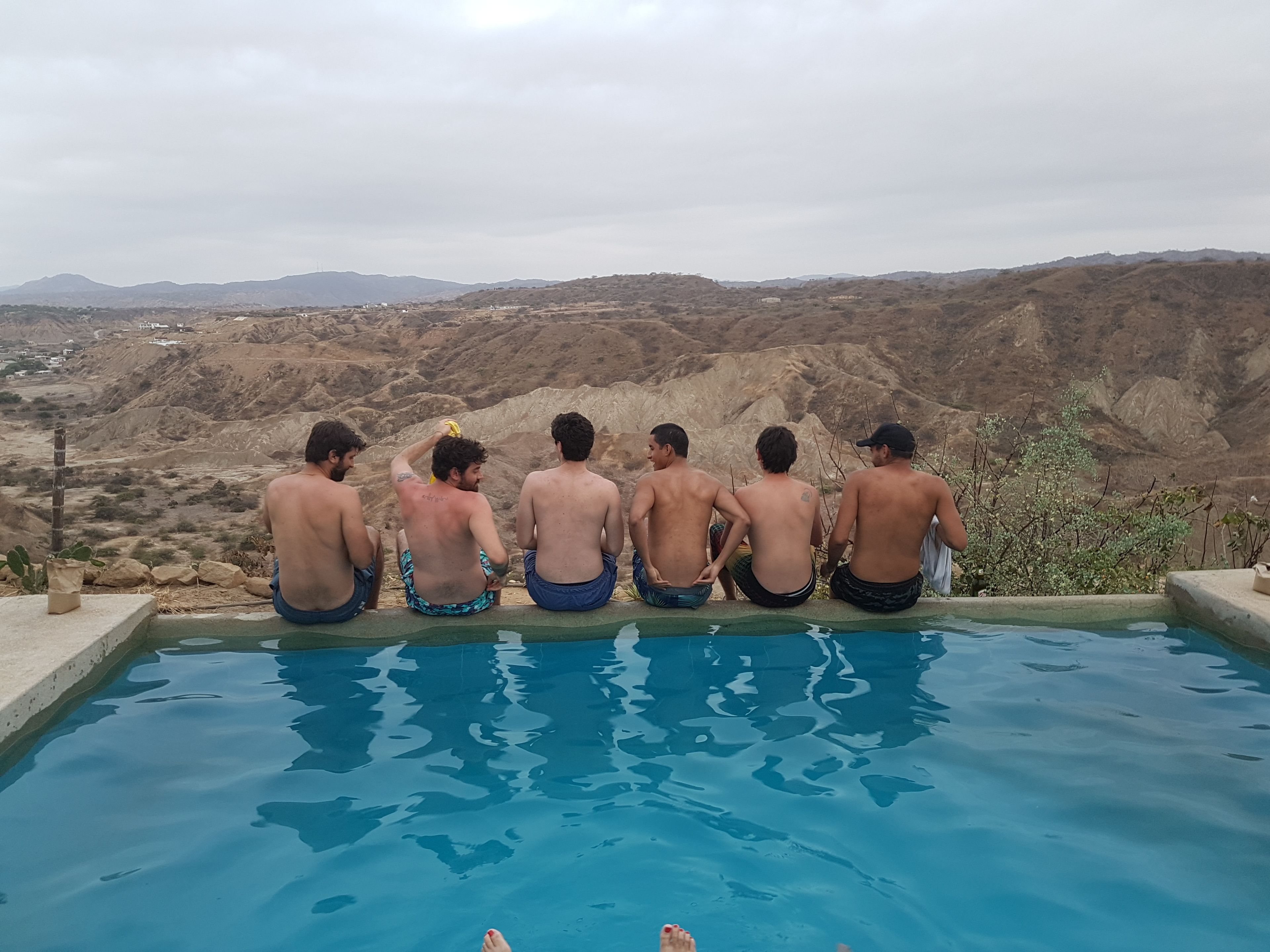 Un grupo de hombres sentados junto a una piscina, con un paisaje montañoso al fondo.