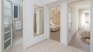 Un pasillo amplio con un espejo grande, una puerta que da a un baño, y un dormitorio con cama y candelabro.