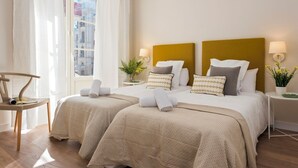Habitación de hotel con dos camas, una silla, un ventanal con cortinas, una planta y una lámpara.